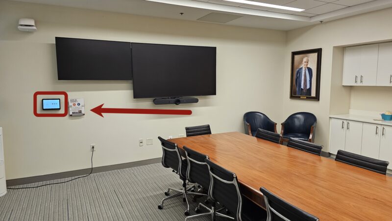Picture of a small conference room in building E52 pointing to the Zoom Room Controller touchpanel on the wall