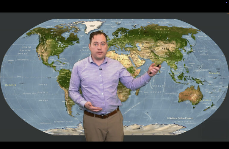 Presenter stands in front of a large world map, gesturing toward Asia while holding a small remote.