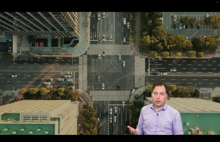 Aerial view of a large city intersection with marked crosswalks, traffic lanes, cars, surrounding buildings, trees, and a presenter speaking in the lower-right corner.