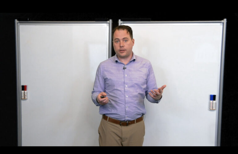Presenter stands between two blank whiteboards, holding a small remote and gesturing while speaking.