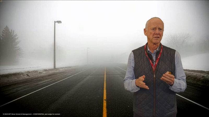Presenter stands in front of a foggy road scene with snow along the shoulders and streetlights fading into the distance. He wears a gray vest over a light blue shirt and holds a small remote while speaking.