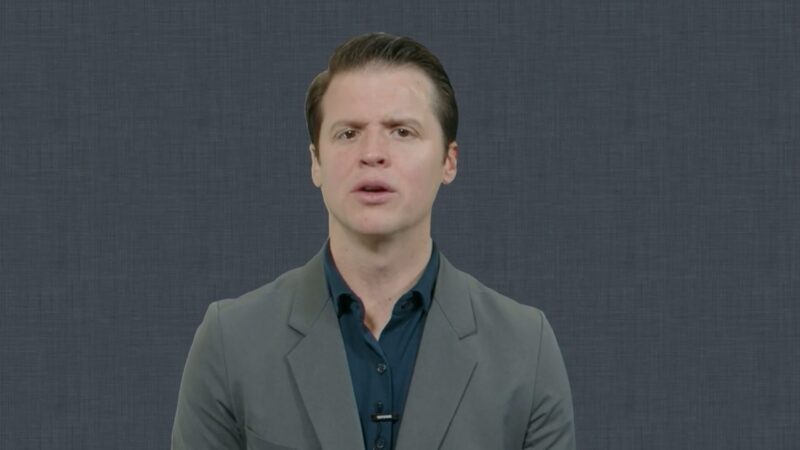 Presenter in a gray blazer and dark blue shirt speaks directly to the camera against a plain textured gray background, wearing a clip-on microphone.